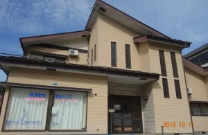 2階建ての介護施設の建物外観。ベージュ色のサイディング仕上げで、玄関入口が見える。