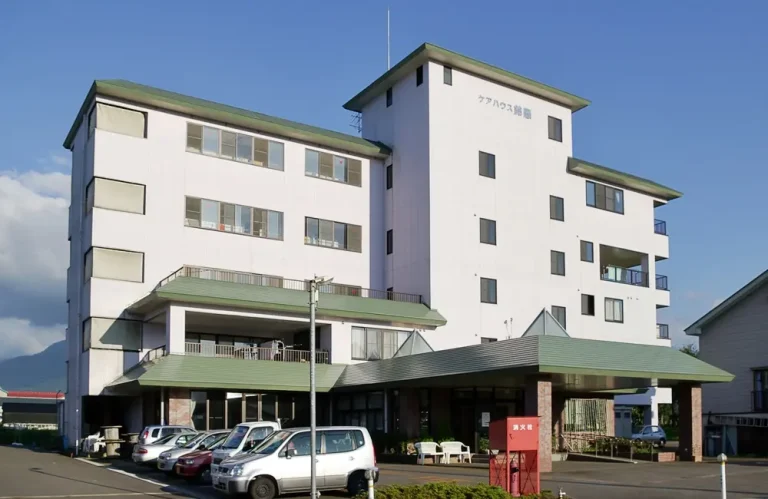 白壁に緑屋根の介護施設。駐車場と玄関前の屋根付きポーチが見える。